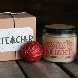 May include: A brown gift box with a white ribbon and a sticker that says "TEACHER" on it. Next to the box is a candle with a label that says "school is Merry & BRIGHT because of you!" There are three red glitter ornaments on the table.