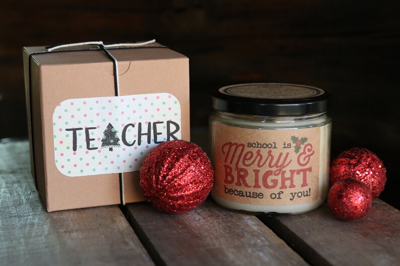 May include: A brown gift box with a white ribbon and a sticker that says "TEACHER" on it. Next to the box is a candle with a label that says "school is Merry & BRIGHT because of you!" There are three red glitter ornaments on the table.