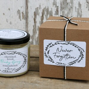 May include: A glass jar candle with a black lid and a white label that reads "cherished. loved. Baby Gray remembered. always." The candle is in a brown box with a white label that reads "Never Forgotten" with a black wreath design.