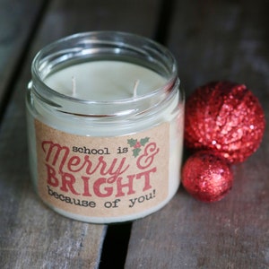 May include: A clear glass jar candle with a brown label that reads "school is Merry & BRIGHT because of you!" The candle is surrounded by two red sparkly ornaments.