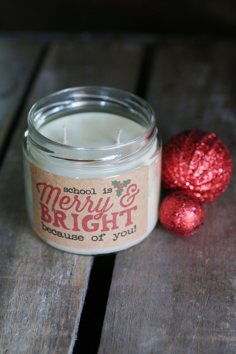 May include: A clear glass jar candle with a brown label that reads "school is Merry & BRIGHT because of you!" The candle is surrounded by two red sparkly ornaments.