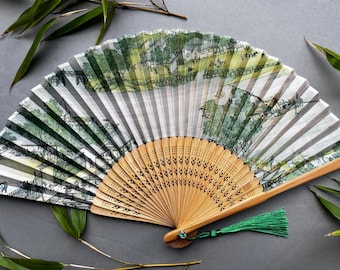 May include: A hand fan with a green and white painted design of a landscape. The fan is made of bamboo and has a green tassel.