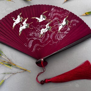 May include: A claret-coloured hand fan with a black frame and a red tassel. The fan depicts white cranes in flight over stylised waves. Bamboo leaves are scattered around the fan.