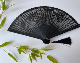 Puede incluir: Un abanico plegable negro con un diseño delicado e intrincado y una borla negra. El abanico está hecho de madera y tiene un estilo japonés tradicional.