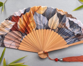 May include: A folding hand fan with a bamboo frame and a leaf pattern in shades of brown, orange, and gray. The fan has a red tassel and is surrounded by green bamboo leaves.