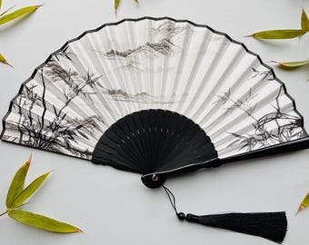 May include: A decorative hand fan with a black handle and a white paper fan. The fan features a black and white landscape design with bamboo and mountains. A black tassel hangs from the base of the fan.