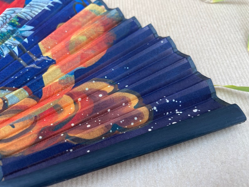 Pu&ograve; includere: Un ventaglio pieghevole blu con un motivo floreale colorato e macchie bianche. Il ventaglio &egrave; chiuso e il manico &egrave; in legno blu scuro.