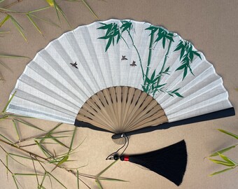 May include: A white folding fan with a black handle and a black tassel. The fan has a green bamboo design with small brown birds.