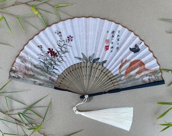 May include: A white folding fan with a black handle and a white tassel. The fan is decorated with a painted scene of flowers, a butterfly, and a rabbit. The fan has Chinese characters written on it.