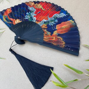Pu&ograve; includere: Un ventaglio pieghevole nero con un design blu, rosso e oro con gru e nuvole. Il ventaglio ha un fiocco nero.