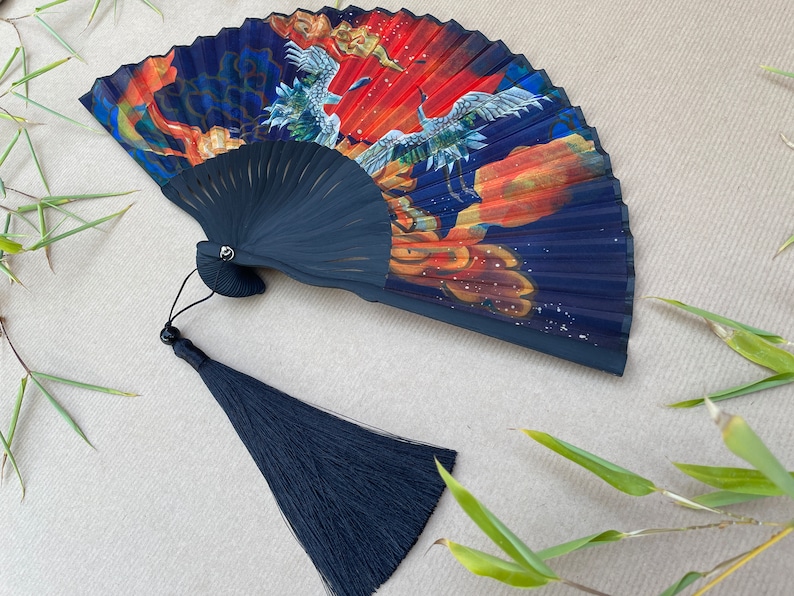 Pu&ograve; includere: Un ventaglio pieghevole nero con un design blu, rosso e oro con gru e nuvole. Il ventaglio ha un fiocco nero.