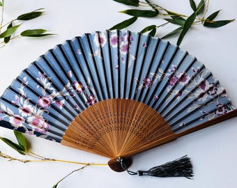 May include: A blue and white hand fan with a floral pattern and a wooden handle. The fan has a black tassel.