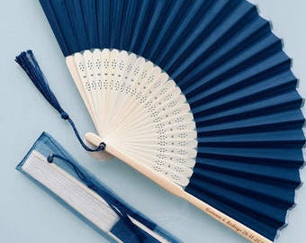 May include: A navy blue silk folding fan with a white wooden handle and a white tassel. The handle is engraved with the text "Autumn & Rodrigo 26.11.2022". The fan is packaged in a blue organza bag.