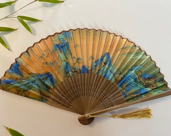 Puede incluir: Un abanico de mano con marco de madera y un panel de papel de seda con un paisaje pintado de montañas y árboles en tonos azules, verdes y amarillos. El abanico tiene un borla dorada.