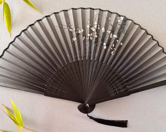 May include: A black and gray hand fan with a floral design. The fan is open, displaying a gradient of gray tones. White flowers and black branches are painted on the fan. A black tassel hangs from the base.