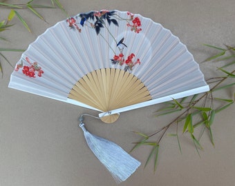 May include: A white folding fan with a floral and bird design. The fan has a wooden handle and a white tassel.