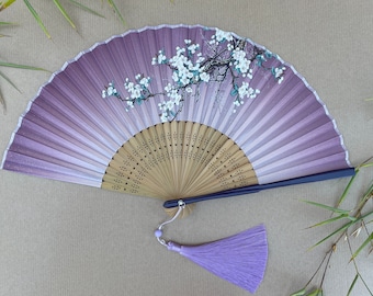 May include: A purple silk folding fan with a white floral design and a wooden handle. The fan has a purple tassel attached to the handle.
