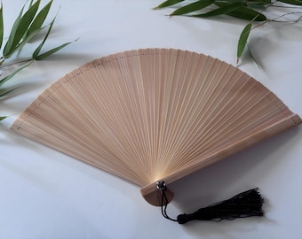 Puede incluir: Un abanico plegable de bambú natural con un borla negra. El abanico está cerrado y tiene un acabado de madera natural.