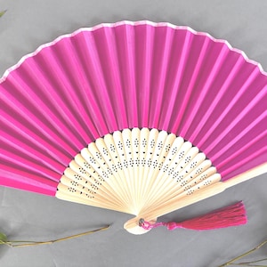 Puede incluir: Un abanico de mano rosa vibrante con un diseño plisado. El abanico presenta un mango de madera beige claro y una borla a juego. El abanico está abierto, mostrando su forma completa, y está sobre un fondo gris con hojas de bambú.