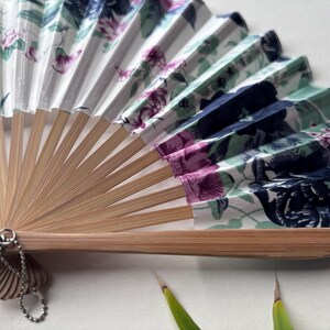 May include: A white folding fan with a floral pattern in shades of pink, blue, and green. The fan is made of bamboo and has a decorative chain attached to the handle.
