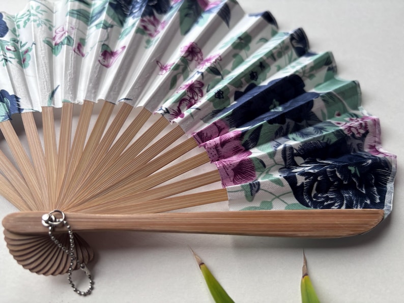 May include: A white folding fan with a floral pattern in shades of pink, blue, and green. The fan is made of bamboo and has a decorative chain attached to the handle.