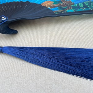 Puede incluir: Un abanico plegable negro con un dise&ntilde;o floral colorido, al que se adjunta una borla de seda azul.