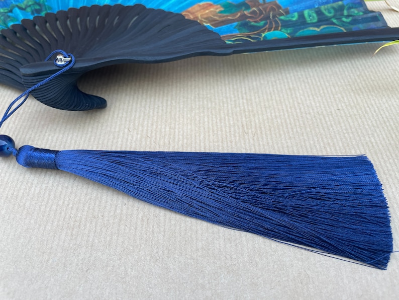 Puede incluir: Un abanico plegable negro con un dise&ntilde;o floral colorido, al que se adjunta una borla de seda azul.