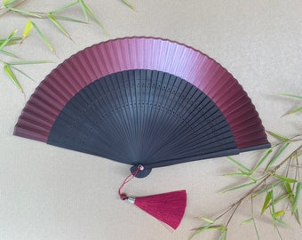 May include: A black and burgundy folding fan with a red tassel. The fan is made of wood and has a delicate design.