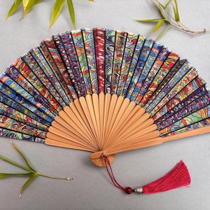 May include: A colorful hand fan with a wooden handle and a vibrant, patterned fabric. The fan features a red tassel and is decorated with a variety of colors, including blue, green, red, and yellow. Bamboo leaves are in the background.