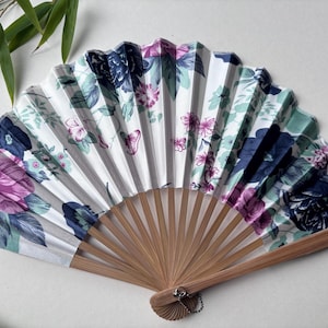 May include: A white folding fan with a floral pattern in shades of pink, blue, and green. The fan has a bamboo handle and a silver tassel.