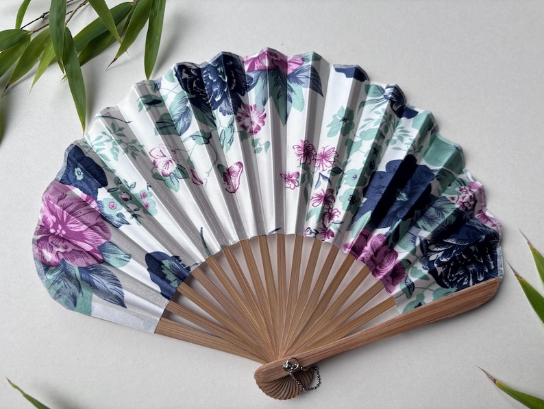 May include: A white folding fan with a floral pattern in shades of pink, blue, and green. The fan has a bamboo handle and a silver tassel.