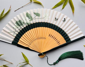 May include: A decorative folding fan with a bamboo frame and a white paper fan. The fan features a green and white floral design with butterflies. A green tassel hangs from the bottom. The fan is open and displayed on a gray surface.