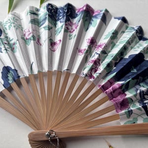 May include: A white folding fan with a floral pattern in shades of pink, blue, and green. The fan is made of fabric and has a bamboo handle.