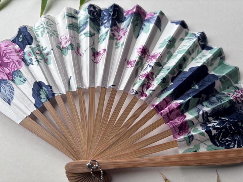 May include: A white folding fan with a floral pattern in shades of pink, blue, and green. The fan is made of fabric and has a bamboo handle.