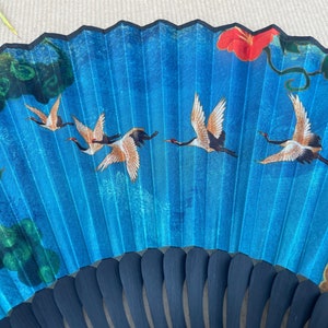 Puede incluir: Un abanico plegable azul con un mango negro y un dise&ntilde;o de p&aacute;jaros volando en un cielo azul con una flor roja y hojas verdes.