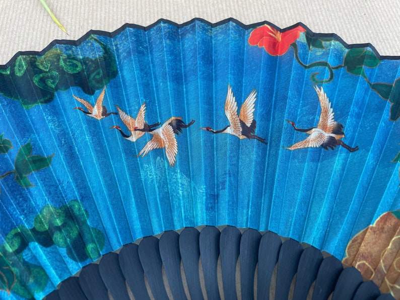 Puede incluir: Un abanico plegable azul con un mango negro y un dise&ntilde;o de p&aacute;jaros volando en un cielo azul con una flor roja y hojas verdes.