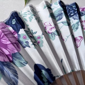 May include: A white folding fan with a floral pattern. The fan features pink, purple, blue, and green flowers on a white background. The fan is made of fabric and has wooden sticks.