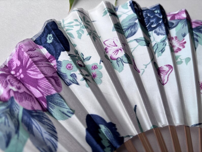 May include: A white folding fan with a floral pattern. The fan features pink, purple, blue, and green flowers on a white background. The fan is made of fabric and has wooden sticks.
