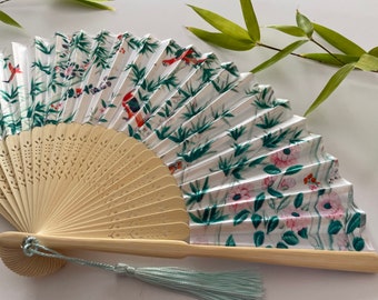 Puede incluir: Un abanico plegable blanco con un diseño floral verde y rosa y un mango de bambú. El abanico tiene un marco de madera tallada y una borla azul claro.
