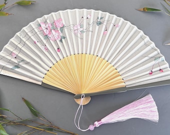 May include: A hand fan with a light gray and white striped design, adorned with pink floral accents and a tassel. The fan's base is made of light wood, and the tassel is pink. The fan is open, displaying its full design.