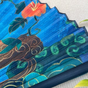 Puede incluir: Un abanico plegable azul con un dise&ntilde;o floral. El abanico presenta una flor roja, hojas verdes y detalles dorados. El abanico est&aacute; hecho de papel y tiene un mango negro.