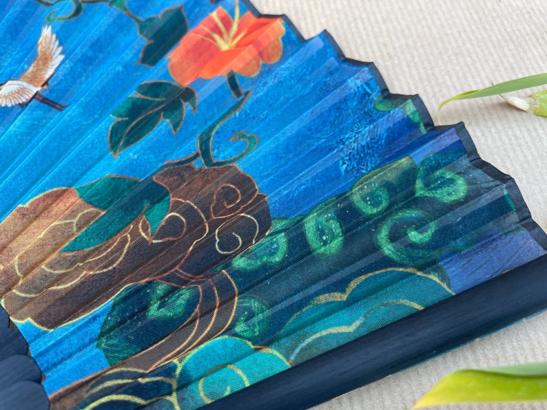 Puede incluir: Un abanico plegable azul con un dise&ntilde;o floral. El abanico presenta una flor roja, hojas verdes y detalles dorados. El abanico est&aacute; hecho de papel y tiene un mango negro.