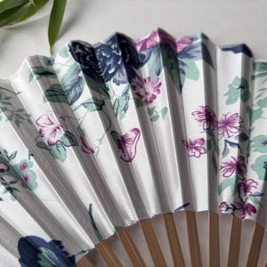 May include: A white folding fan with a floral pattern in shades of pink, blue, and green. The fan is made of paper and has a wooden handle.