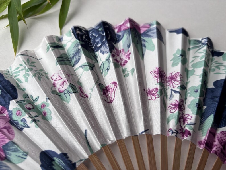 May include: A white folding fan with a floral pattern in shades of pink, blue, and green. The fan is made of paper and has a wooden handle.