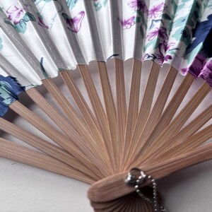 May include: A white folding fan with a floral pattern in shades of blue, green, and purple. The fan is made of bamboo and has a silver chain attached to the handle.
