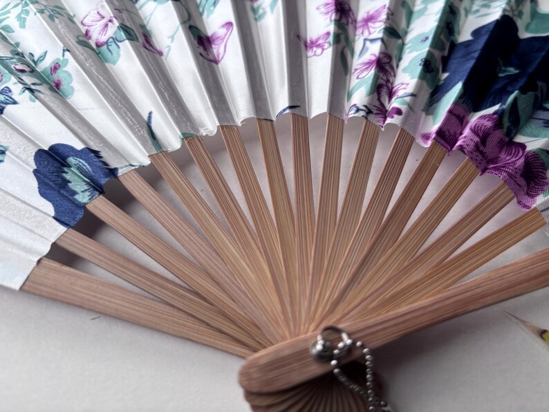 May include: A white folding fan with a floral pattern in shades of blue, green, and purple. The fan is made of bamboo and has a silver chain attached to the handle.