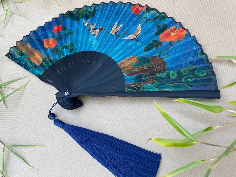Puede incluir: Un abanico azul y negro con un dise&ntilde;o floral y de p&aacute;jaros. El abanico tiene un borla azul.
