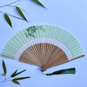 Op de afbeelding: Een vouwbare handwaaier met een lichtgroen stoffen blad voorzien van een delicate illustratie van bamboebladeren. De waaier heeft een bruin houten frame en een donkergroene decoratieve kwast aan de basis, versierd met een kleine zwarte kraal.