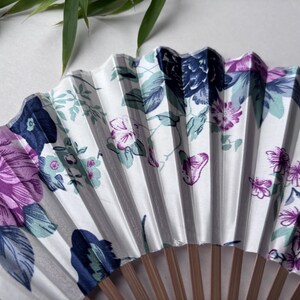 May include: A white folding fan with a floral design featuring purple, blue, and green flowers. The fan is made of paper and has a wooden handle.