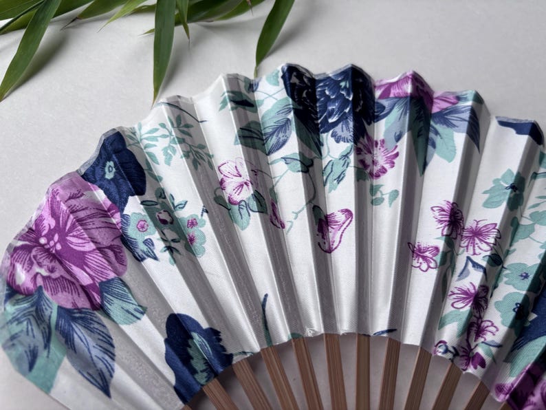 May include: A white folding fan with a floral design featuring purple, blue, and green flowers. The fan is made of paper and has a wooden handle.